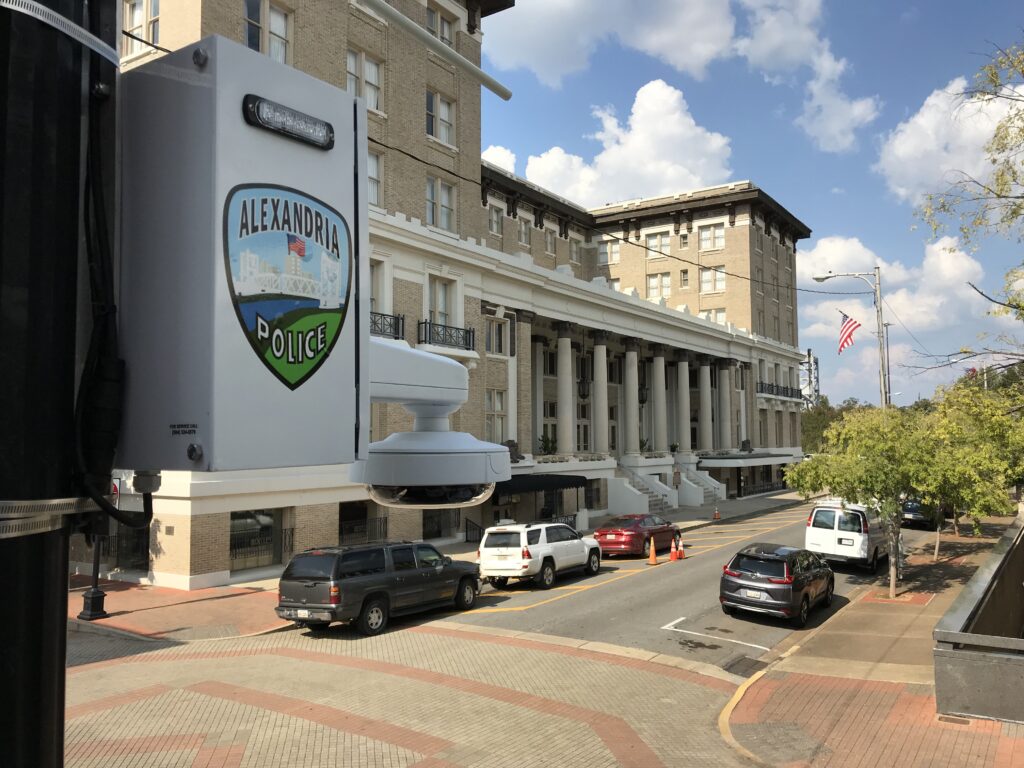 A street view featuring an Alexandria Police sign, vehicles on the road, and a large building with columns and balconies under a partly cloudy sky.